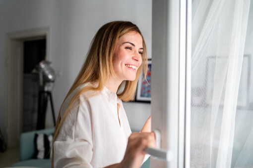 AdobeStock_377893241 Beautiful smiling young woman opening window at the morning, fresh air,